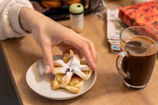 A woman picks freshly baked fruit puff pastry on the ceramic plate next to hot cup of coffee