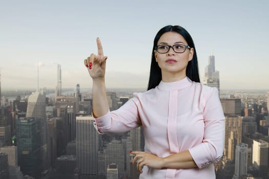 Young asian woman touching invisible virtual screen with her finger. Urban cityscape background.
