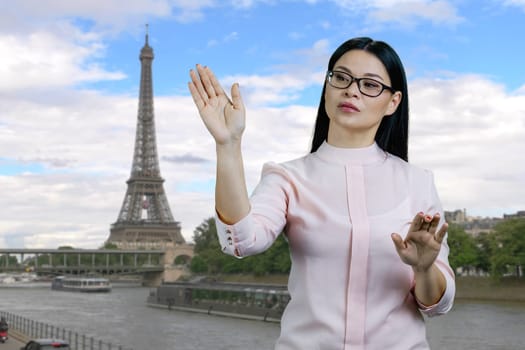 Young asian business woman swipes invisible virtual screen standing outdoors. France Eiffel Tower background.