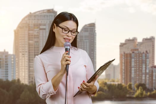 Young asian businesswoman with microphone is reading from clipboard. Outdoors with urban scape background.