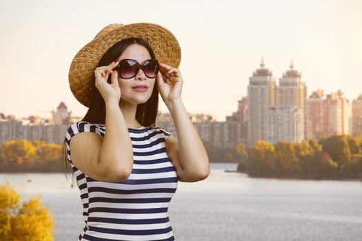 Young brunette woman puts on sunglasses outdoors. City river background.