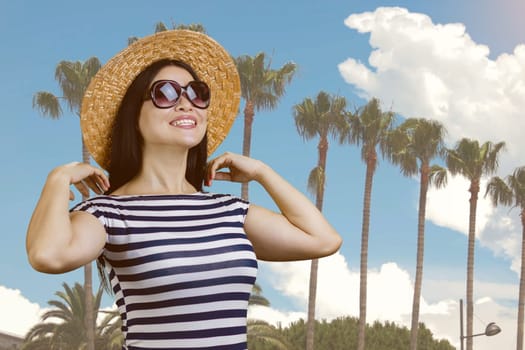Young smiling woman with straw hat standing outdoors. Palm trees and blue sky background.