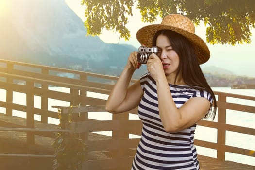 Young tourist woman in straw hat taking a photo on retro photo camera. Summer resort landscape with sunshine.