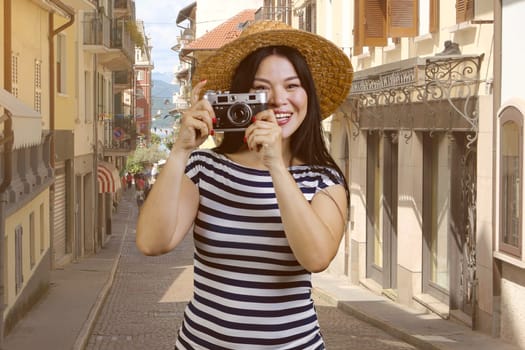 Portrait of a cheerful asian woman in straw hat holding vintage photo camera. European street background.