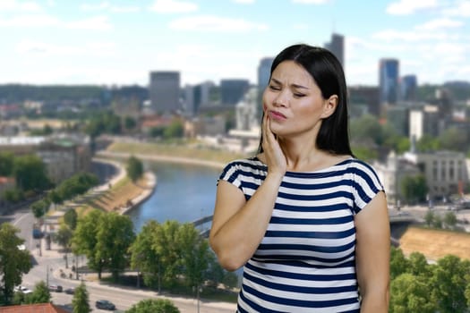 Young asian woman touching her cheek because of toothache. Urban cityscape background.
