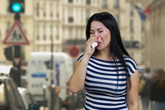 Asian woman with runny nose standing outdoors on the street. Blurred urban scape background.