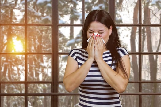 Asian woman with runny nose standing indoors. Sneezing brunette on checkered windows background with winter evening landscape.
