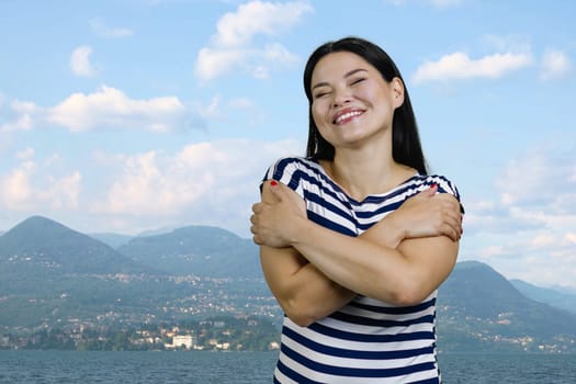Happy young asian woman is hugging herselft outdoors. Mountain scape background.