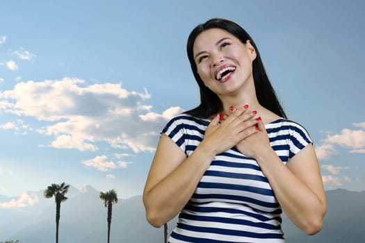 Young asian brunette woman is laughing hard standing outdoors. Blue sky background.