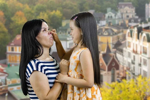 Mom and her little girl are fooling around together outdoors. Making a piggy nose. European town scape background.