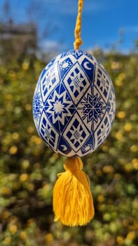 A striking Ukrainian pysanka hangs against a backdrop of blooming flowers, showcasing vibrant blue patterns and a golden tassel.