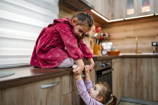 Two young sisters display their bond in a cozy kitchen setting. One girl sits joyfully on the counter, while the other playfully tries to pull her down, creating a tender and lively atmosphere.