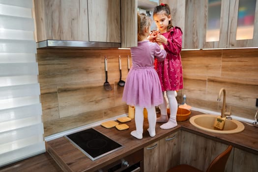 In a stylish kitchen bathed in sunlight, two little girls engage in a delightful playtime, one standing on a counter while the other carefully hands her ingredients.