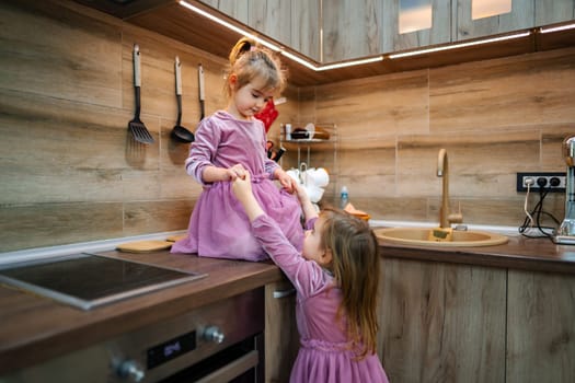 Two young sisters engage in playful antics within a warm, modern kitchen, showcasing joyous expressions as they share a light-hearted moment surrounded by kitchenware.