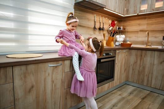 In a cozy and stylish kitchen, two young girls are immersed in playful fun, one perched on the counter while the other reaches up joyfully, fostering creativity and laughter in their enchanting world.