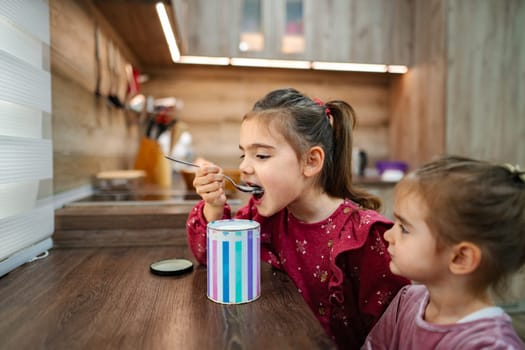 In a warm, inviting kitchen, two sisters share a delightful moment as one eagerly tastes a treat from a colorful container. The atmosphere is filled with joy and laughter, highlighting their bond.