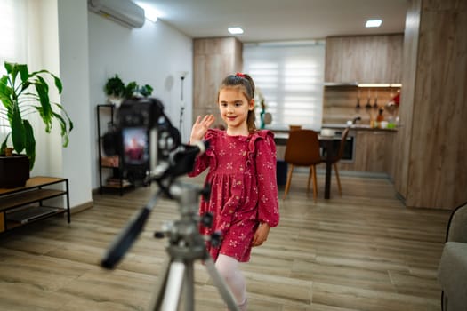 A cheerful young girl wearing a dress smiles and waves while standing in a contemporary kitchen. Bright sunlight spills in, enhancing her playful spirit as she engages with the camera.