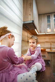 Two cheerful young girls in matching dresses engage in playful fun in a warm, inviting kitchen filled with natural light. Their laughter fills the air as they bond over imaginative games.