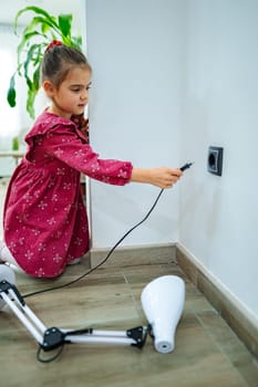 Curious and focused, a young girl in a pink dress kneels beside the wall to connect a robotic arm to a charging outlet in a modern living space.