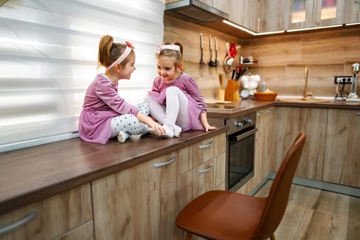 Two cheerful girls dressed in matching outfits sit on a stylish kitchen counter, sharing laughter and playful moments. Sunlight pours in, creating a warm atmosphere filled with joy.
