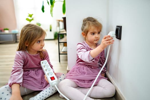 In a cozy living space, two young girls sit on the floor, one curiously reaching for an electrical outlet while the other observes.