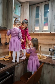 Three young girls, dressed in matching purple dresses, playfully explore a modern kitchen, reaching for ingredients as they prepare a snack, creating joyful laughter and cherished memories.