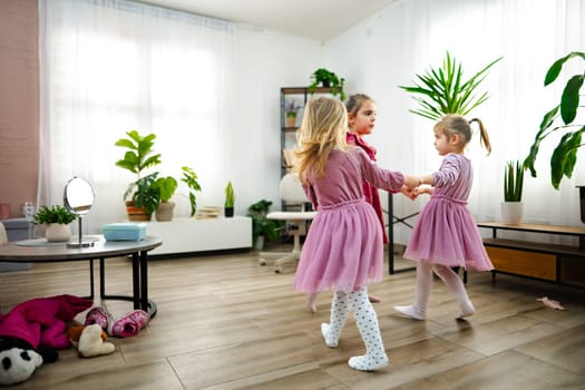 Three young girls in pink dresses dance joyfully in a sunlit living room. Surrounded by plants and soft toys, they hold hands while spinning, creating a lively and heartwarming atmosphere.