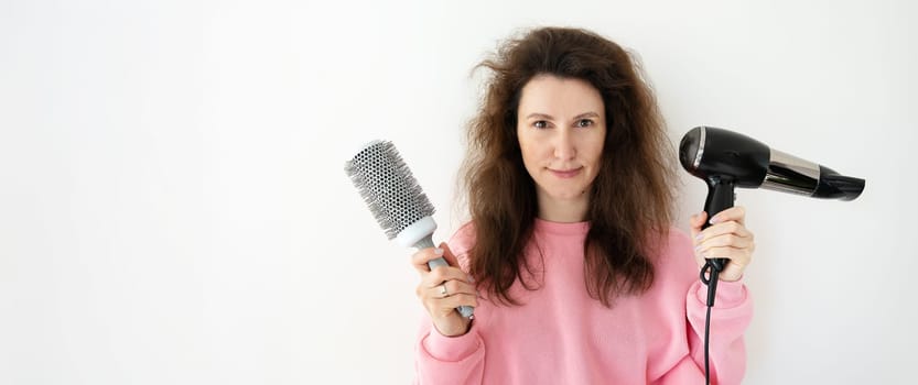 A woman with messy hair holds a round hairbrush and hairdryer, preparing to style her hair.