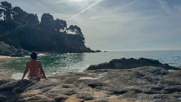 A woman in a pink bikini sits on the sandy beach of Lloret de Mar, Spain. Her face is not visible, focusing on the relaxed atmosphere of the beach and the beautiful surroundings. Space for text.
