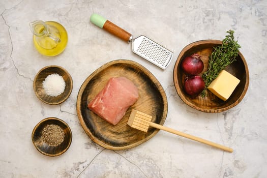 Freshly sourced pork chop is laid on a wooden plate alongside red onions, thyme, cheese, and seasoning. Cooking tools like a grater and a wooden mallet are nearby, ready for meal prep.