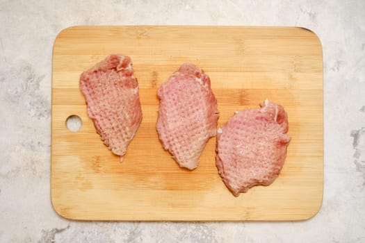 Three raw pork chop cuts are on a wooden chopping board. The fresh meat is awaiting seasoning before being cooked on a stovetop or grill for a delicious meal.