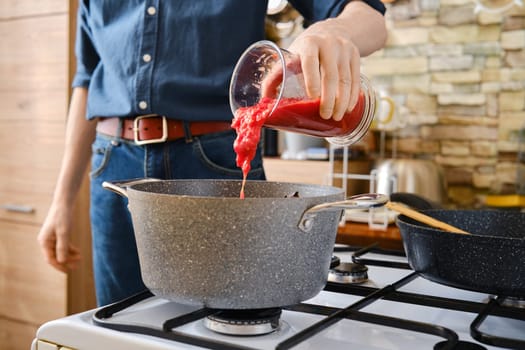 A person pours tomato sauce from a jar into a pot on the stove. The kitchen features modern decor with a warm ambiance.