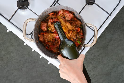 A hand adds a bottle of wine to a pot filled with meat and vegetables simmering on the stove. The cozy kitchen features a gas burner and a modern design, perfect for creating delicious meals.