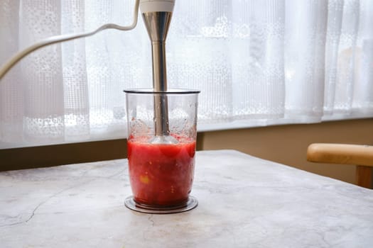 Bright red tomatoes are being blended in a clear glass container using an immersion blender. The kitchen features a bright atmosphere with natural light filtering through sheer curtains.