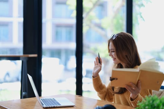 Smiling young woman engaged in a joyful online e-learning video call, gesturing while holding a book with headphones on a wooden table nearby.