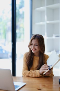 Smiling young woman engaged in a joyful online e-learning video call, gesturing while holding a book with headphones on a wooden table nearby.