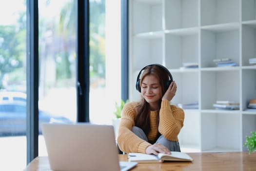 Young woman enjoying a relaxing music break at her desk while using headphones, writing in a notebook, and working on her laptop for calm inspiration.