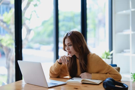 Smiling young woman engaged in a joyful online e-learning video call, gesturing while holding a book with headphones on a wooden table nearby.