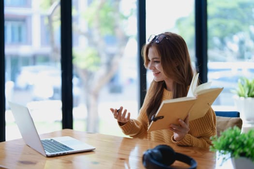 Smiling young woman engaged in a joyful online e-learning video call, gesturing while holding a book with headphones on a wooden table nearby.