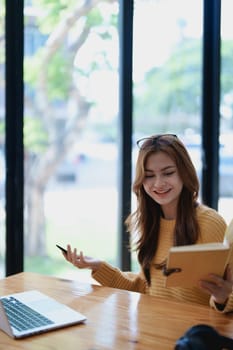 Smiling young woman engaged in a joyful online e-learning video call, gesturing while holding a book with headphones on a wooden table nearby.
