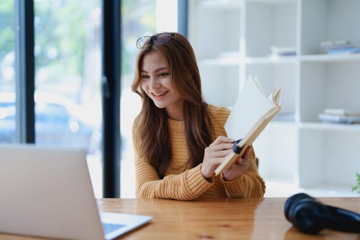 Smiling young woman engaged in a joyful online e-learning video call, gesturing while holding a book with headphones on a wooden table nearby.
