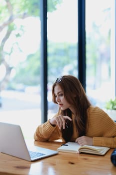 Smiling young woman engaged in a joyful online e-learning video call, gesturing while holding a book with headphones on a wooden table nearby.