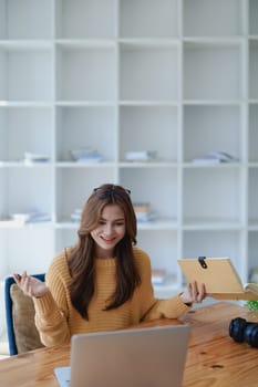 Smiling young woman engaged in a joyful online e-learning video call, gesturing while holding a book with headphones on a wooden table nearby.