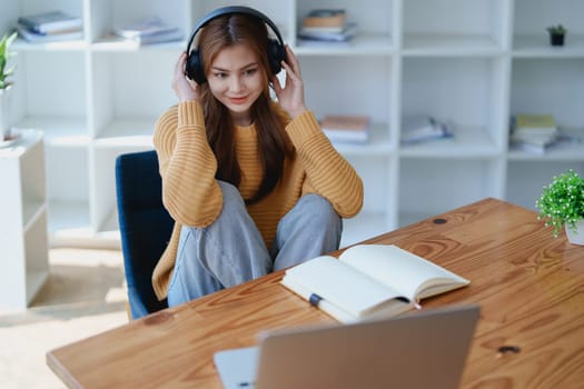Young woman enjoying a relaxing music break at her desk while using headphones, writing in a notebook, and working on her laptop for calm inspiration.