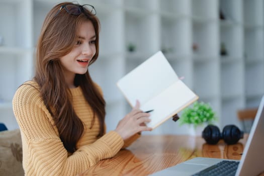 Smiling young woman engaged in a joyful online e-learning video call, gesturing while holding a book with headphones on a wooden table nearby.