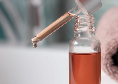 Drop falls from a pipette resting diagonally on a bottle filled with brown liquid against greenbathroom, close-up. Natural serum, gel, oil or other cosmetic product for everyday beauty routine