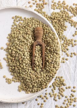 Plate full of dried raw green lentils with a wooden scoop on white table top view. Healthy raw vegetarian food, legumes plant-based protein source. Traditional mediterranean ingredient
