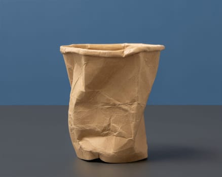 Used and crumpled disposable beige kraft paper cup on grey against blue closeup, negative space. Consumerism concept in modn world. One-use tableware