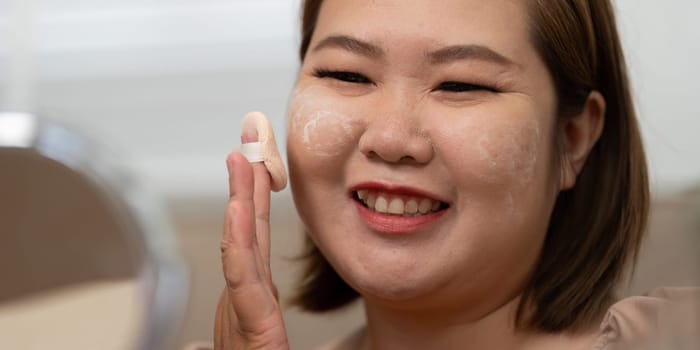 With a bright smile, a woman applies makeup powder, reflecting her joy and confidence as she enhances her beauty in a comfortable environment.