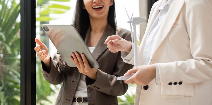 Two professional women engaged in a dynamic discussion about sustainable practices, featuring a small wind turbine as a symbol of renewable energy in an eco-friendly setting.